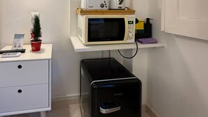 A kitchenette with a microwave, toaster, and coffee maker on a white shelf, a small potted plant, a black trash bin, and a white desk with a keyboard and a small sign.