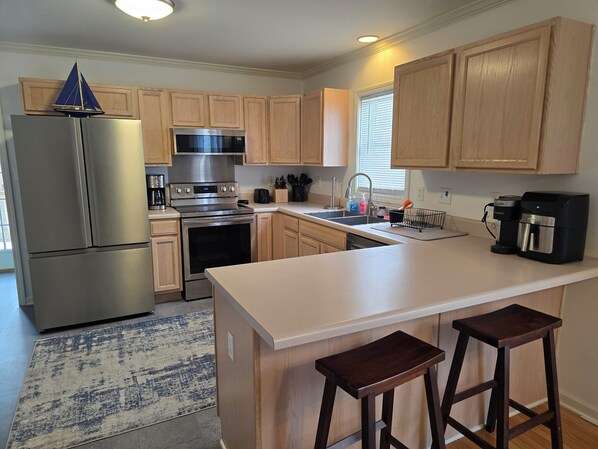 A modern kitchen with wooden cabinets, stainless steel appliances, a large island with bar stools, and a window with blinds. - Beautifully Furnished Townhouse on James Island, 3 minutes to Downtown. (Charleston)