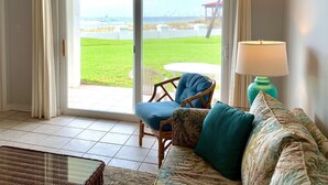 A room with a sliding glass door leading to a patio with a view of the sea and a gazebo. There is a wicker chair and a sofa with a patterned throw pillow. A lamp is on a side table.