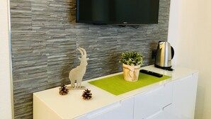 Une cuisine moderne avec un pare-feu en pierre, un téléviseur à écran plat fixé au mur, un meuble blanc, un set de table vert, une machine à café, ainsi que des pommes de pin décoratives et une figurine de chèvre.