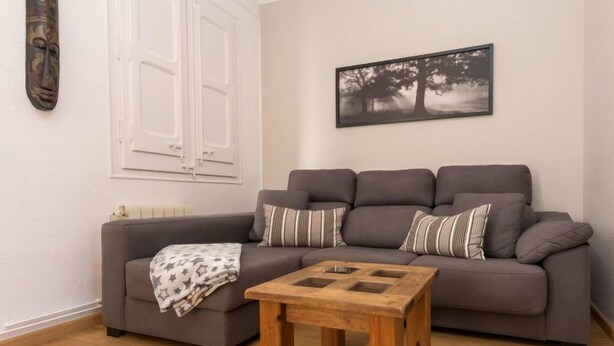 Una sala de estar con un sofá gris, almohadones a rayas, una mesa de centro de madera, un cuadro con árboles y una máscara decorativa en la pared.