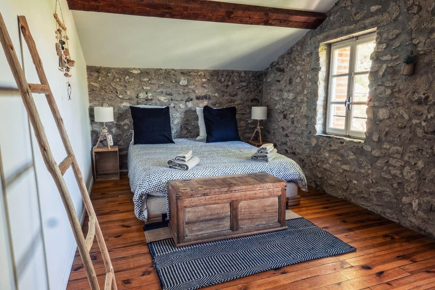 A bedroom with a stone wall, wooden floor, a bed with a checkered bedspread, two pillows, a wooden bench, a nightstand with a lamp, and a window with a view.