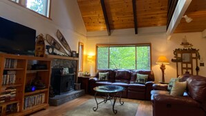 A spacious living room with a high ceiling, wooden beams, a fireplace, a television, and a large window offering a view of trees.