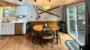 A wooden interior with a dining table, chairs, a kitchen area, and a large window.