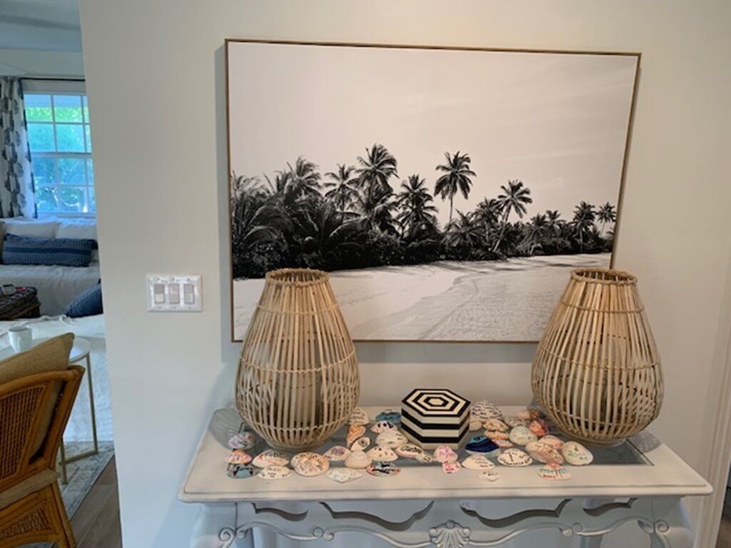 Een kamer met een witte muur, een schilderij van een strandgezicht in een lijst, twee rieten vazen, een decoratief dienblad met schelpen en een rieten stoel.