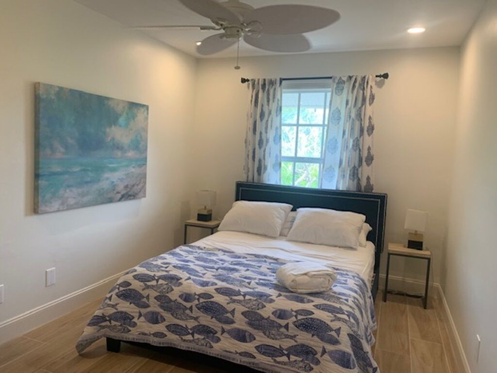 Een slaapkamer met een bed, een plafondventilator, een raam met gordijnen, een schilderij aan de muur en een nachtkastje met een lamp.