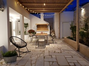 An outdoor patio with a stone floor, a wooden table with chairs, a barbecue grill, and string lights. - Modern House near Parikia with 3 bedrooms & 2 bath (Πάρος)