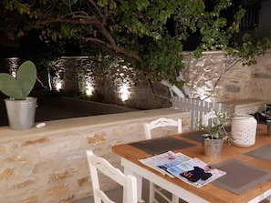 An outdoor seating area with a stone wall, white chairs, a wooden table, and a potted plant. - Modern House near Parikia with 3 bedrooms & 2 bath (Πάρος)