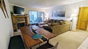 Een woonkamer met een houten eettafel, een bank, een open haard, een televisie en een groot raam.