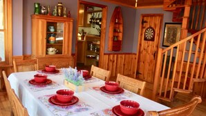 Uma área de refeições com mesa de madeira posta para seis pessoas, rodeada por cadeiras de madeira. Um armário com portas de vidro é visível ao fundo, e uma escada de madeira está à direita.
