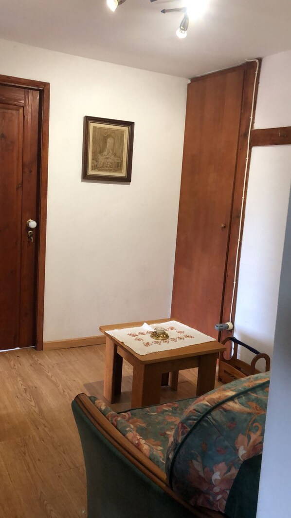 Une pièce avec une porte en bois, un tableau encadré au mur, une petite table recouverte d’une nappe à motifs et un canapé à fleurs.
