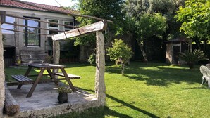 Uma área de estar ao ar livre rústica, com uma mesa e bancos de madeira, uma estrutura de pedra e um pequeno jardim com uma árvore e um vaso de planta.