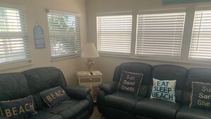 A living room with two black leather sofas, each with a pillow that reads "Sun Sand Shells" and "Eat Sleep Beach." There is a small table with a lamp and a decorative item on the wall.