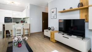 Ein modernes Wohnzimmer mit Flachbildfernseher, Holztisch und Essbereich mit weißen Schränken sowie einer Küche im Hintergrund.