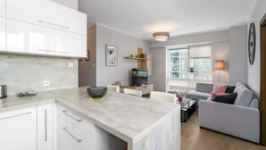 Eine moderne Küche mit weißen Schränken, einer Marmorarbeitsplatte und einem Essbereich mit grauem Sofa und Fernseher.