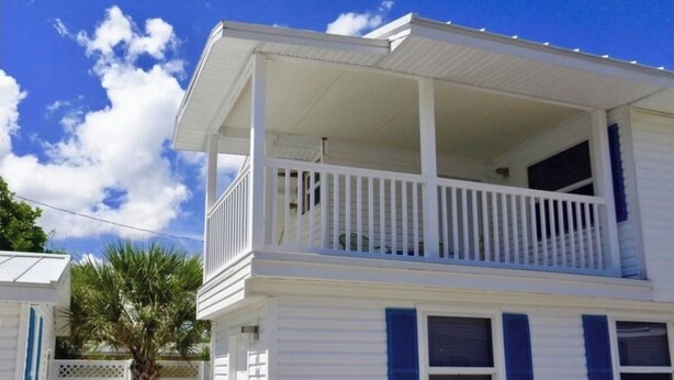 A two-story white house with a balcony, a palm tree, and a white fence.