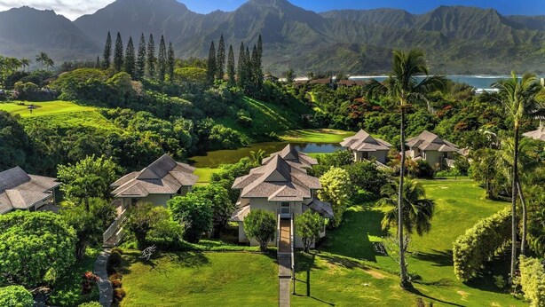 A resort with multiple bungalow-style buildings, a golf course, and a view of a mountain range and ocean.