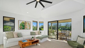A modern living room with a white sofa, a wooden coffee table, and a green chair. There are large windows with a view of greenery and a deck area.