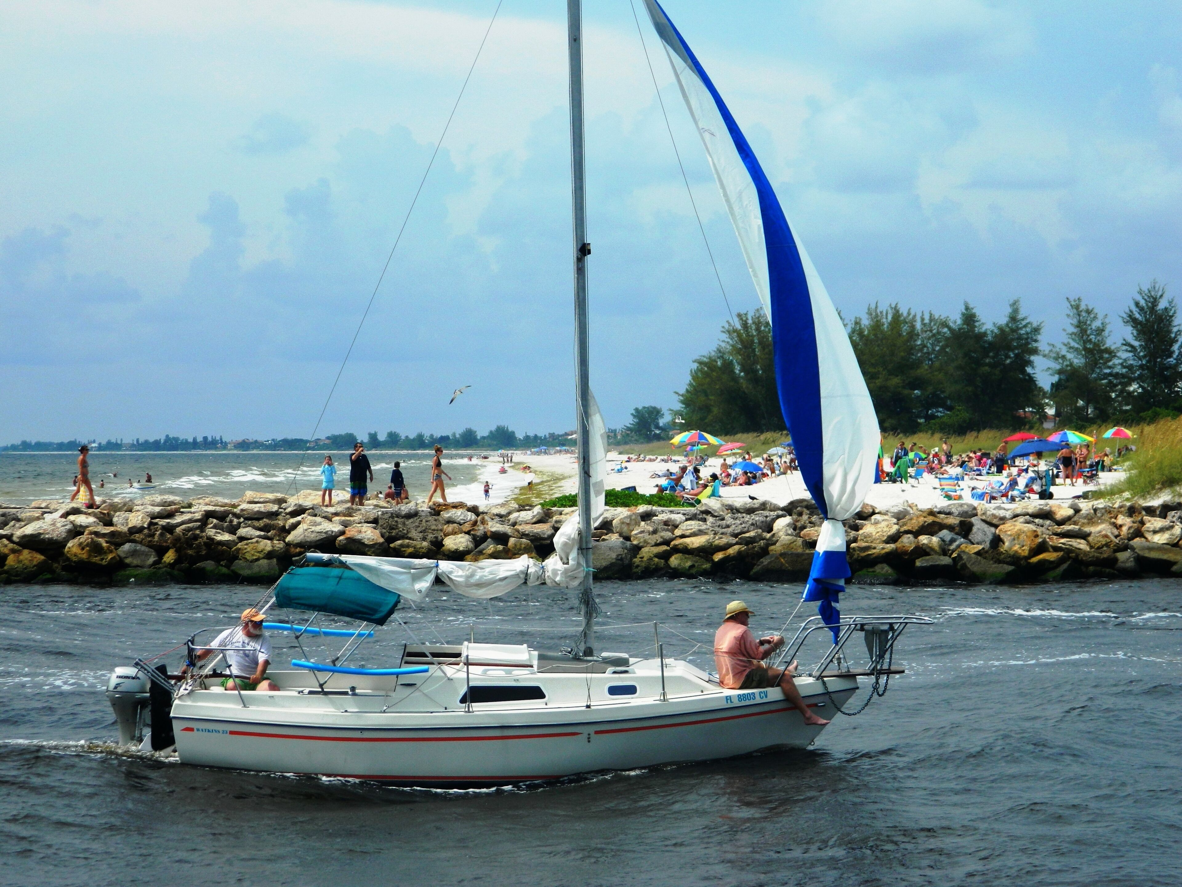 A sailboat with a single mast and a cabin, moving through a body of water near a rocky shore with people on the beach and umbrellas in the background.