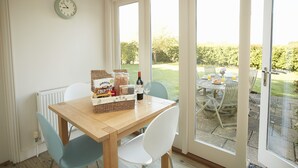 Un coin repas avec une table en bois, des chaises blanches et une vue sur un jardin, visible à travers de grandes fenêtres.