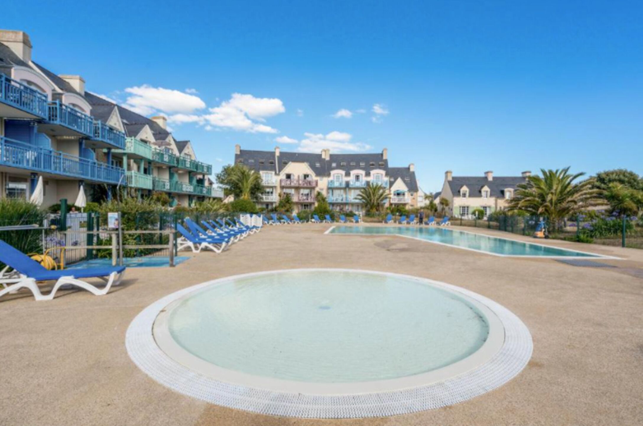 Un complex turístic amb una gran piscina, diversos balcons amb marc blau i un cel serè.