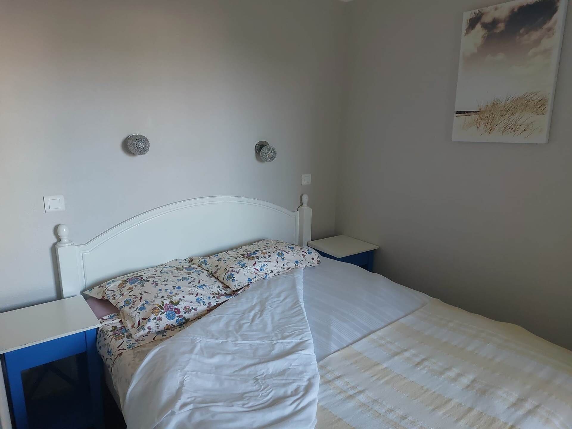 Quarto com cama branca, almofada florida e parede lisa com um pequeno quadro.