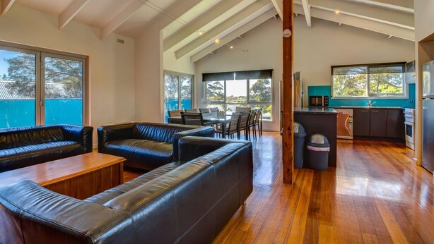 A modern living room with leather sofas, a wooden coffee table, and a dining area with chairs and a table. There is a kitchen with a microwave and a refrigerator. - Raywood - sleeps up to 17 (COWES)