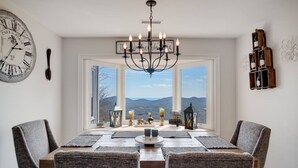 A dining room with a large table, four chairs, a chandelier, a clock, and a view of mountains through large windows. - High Heaven Mountain top dream (Sapphire)