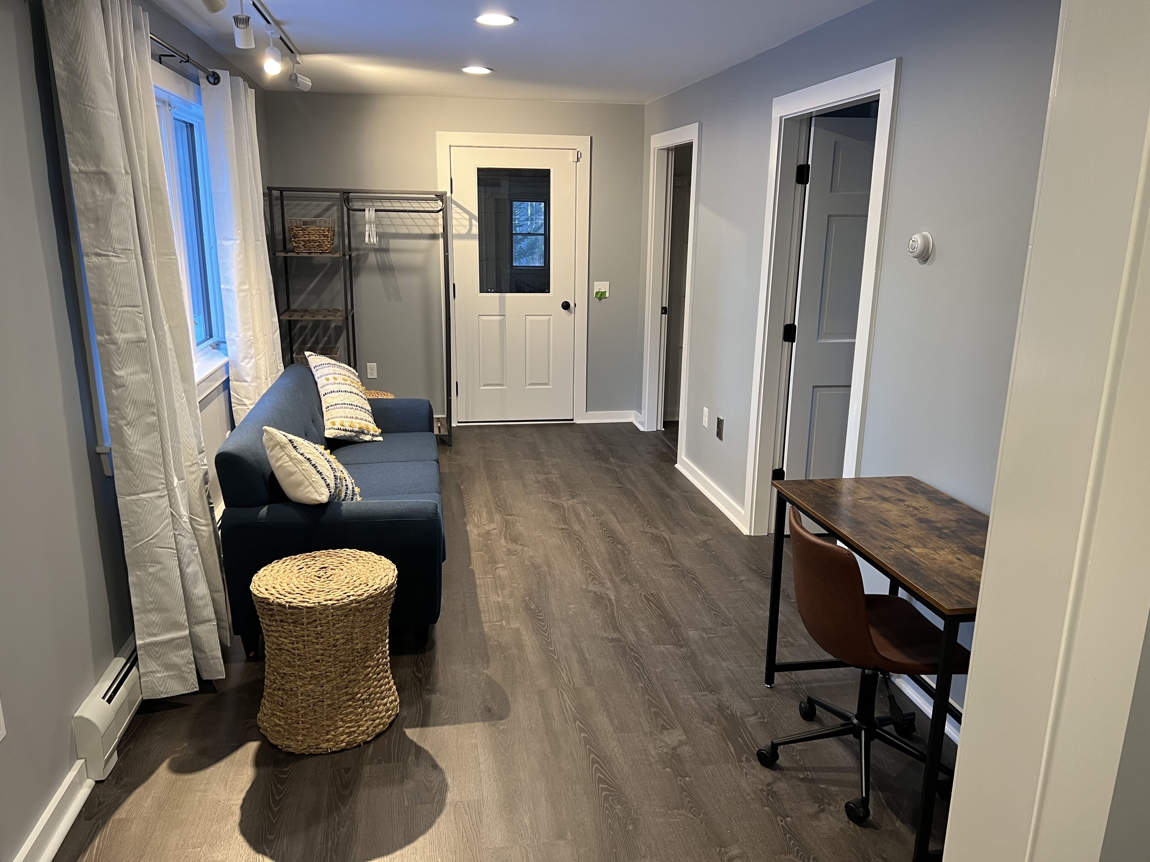 A hallway with a dark blue sofa, wicker ottoman, and a wooden table with a chair.