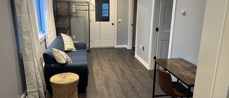 A hallway with a dark blue sofa, wicker ottoman, and a wooden table with a chair.