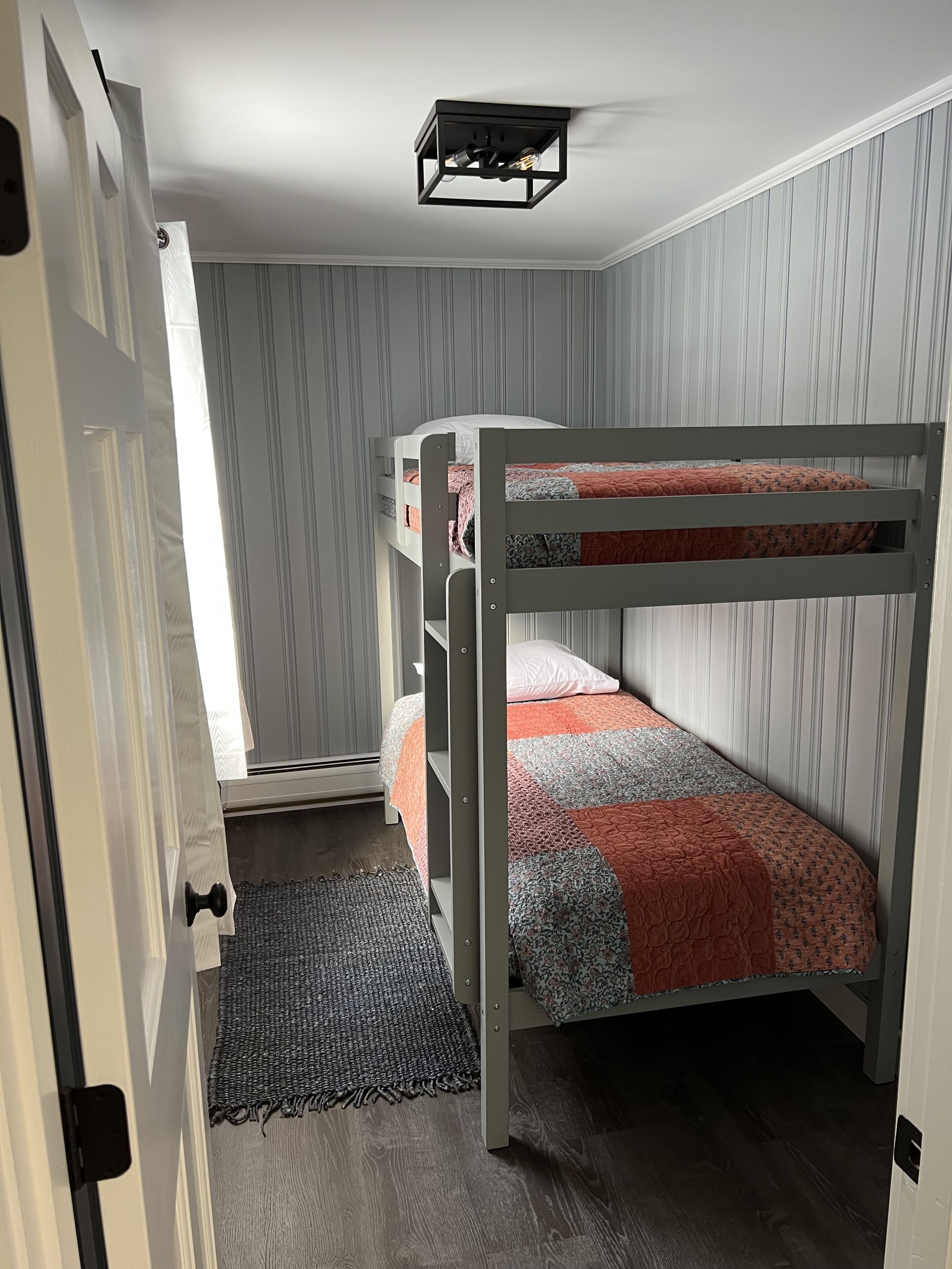 A bunk bed room with a gray and white striped wallpaper, a woven rug, and a ceiling light fixture.