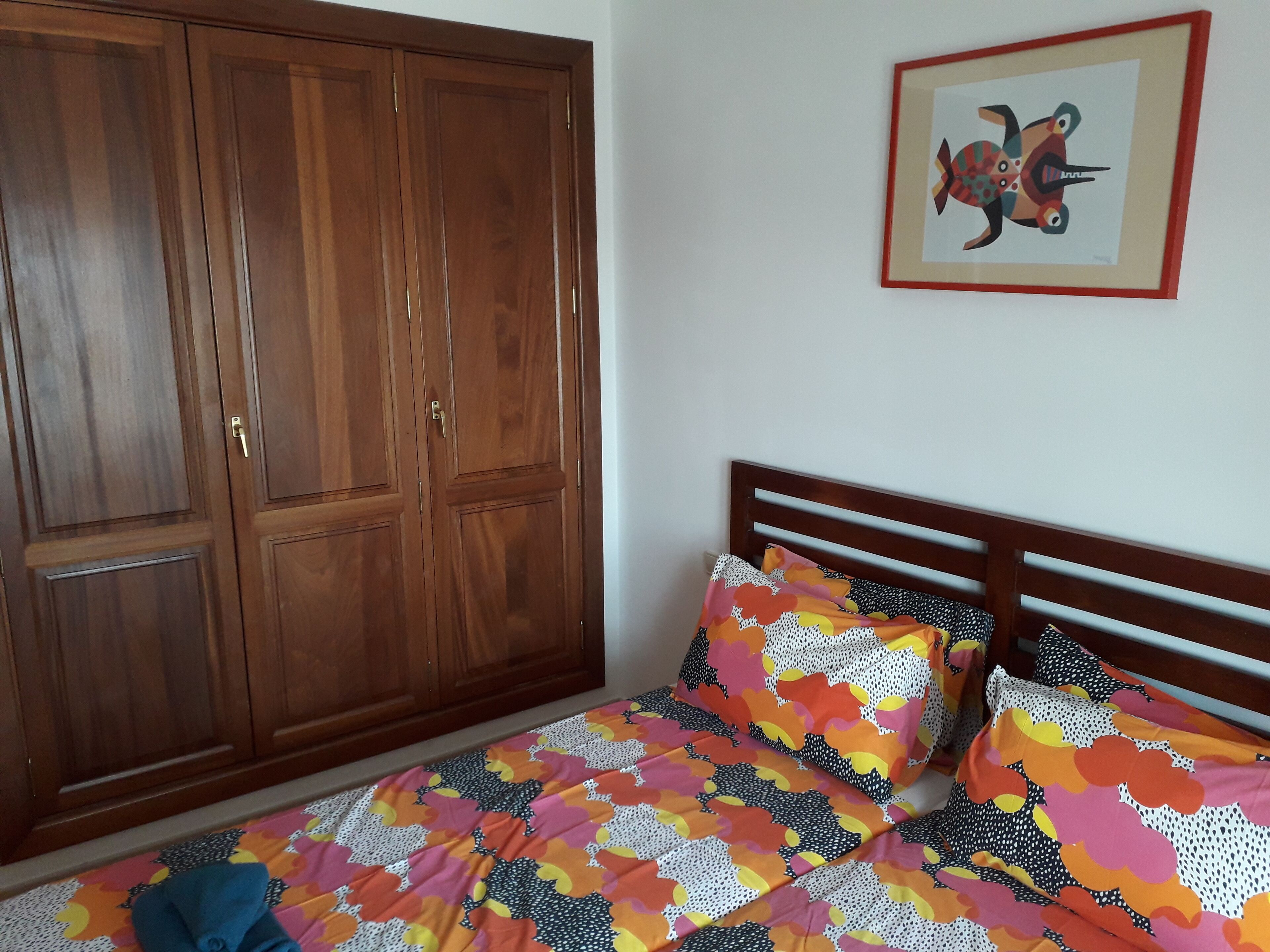 Un dormitorio con un armario de madera, una cama con ropa de cama colorida y un cuadro en la pared.