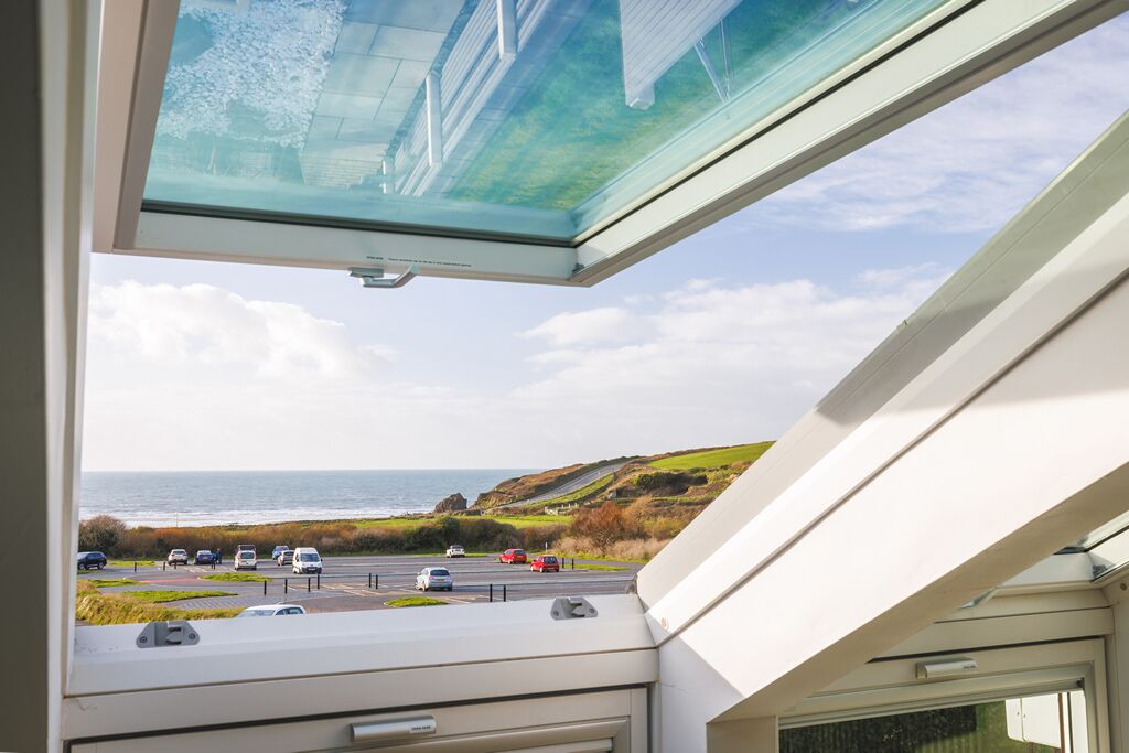 Une vue depuis une fenêtre surmontée d’une lucarne, donnant sur un parking et la mer.