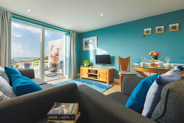 A living room with a sofa, a television, a wooden cabinet, and a dining table with chairs. - Beach View - "cracking sea views" (Broad Haven)