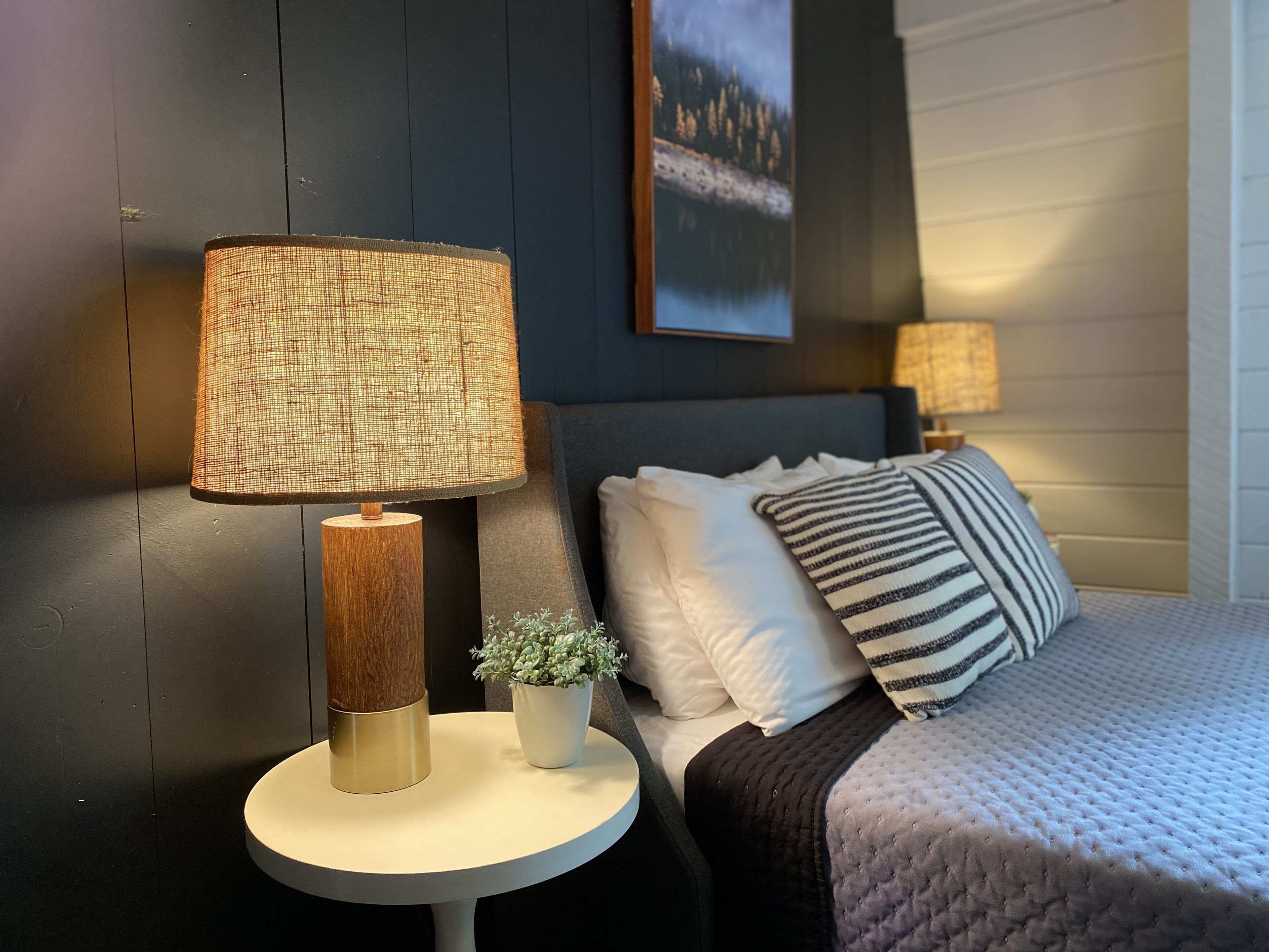 A bedroom with a bed, two bedside lamps, a small potted plant, and a framed picture on the wall.