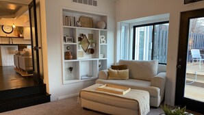 Ein Wohnzimmer mit einem beigefarbenen Sofa, einem Couchtisch und einem Bücherregal. Ein Fenster bietet einen Blick ins Freie.