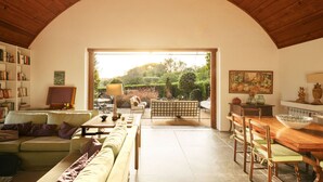 A spacious living area with a high arched ceiling, a sofa set, a dining table, and a view of a garden through an open doorway.
