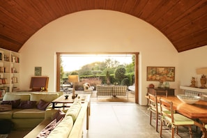 A spacious living area with a high arched ceiling, a sofa set, a dining table, and a view of a garden through an open doorway.