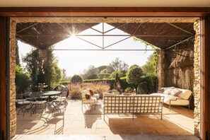 An outdoor patio with a stone wall, a wooden pergola, a dining table with chairs, a sofa, and a bench.