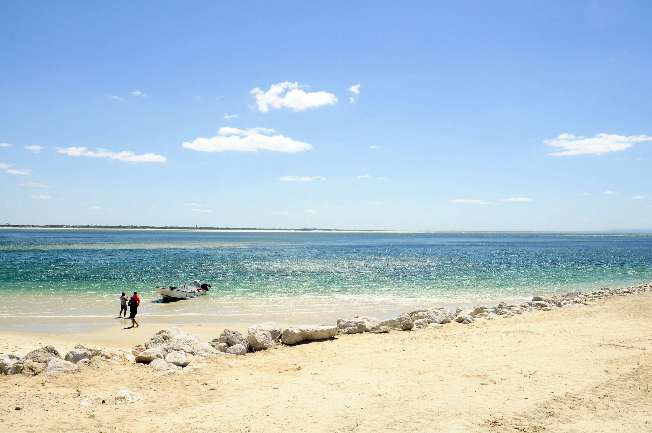 A beach with clear blue water, a rocky shoreline, and a few people walking along the shore.