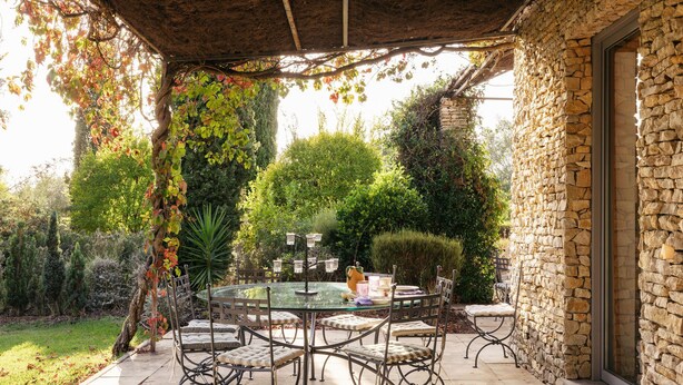 A patio with a round glass table and metal chairs, surrounded by stone walls and greenery.