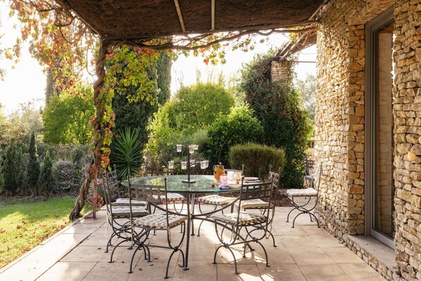 A patio with a round glass table and metal chairs, surrounded by stone walls and greenery.