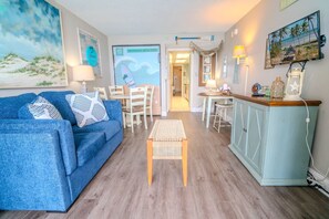 A living room with a blue sofa, a small wooden table, a kitchen area with a light blue cabinet, and a TV displaying a beach scene.