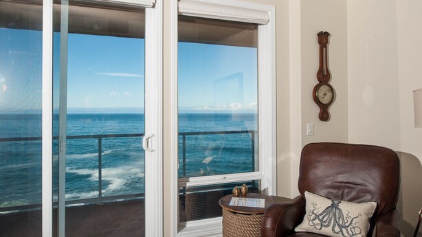 A room with a view of the ocean through a sliding glass door, a brown leather chair with a decorative pillow, and a wall-mounted clock.