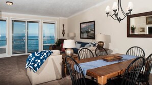 A dining area with a wooden table, chairs, a chandelier, and a view of the ocean through large windows.