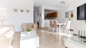 Ein modernes Wohnzimmer mit einem beigen Sofa, einem weißen Couchtisch und einem Essbereich mit weißen Stühlen und einem Glastisch.