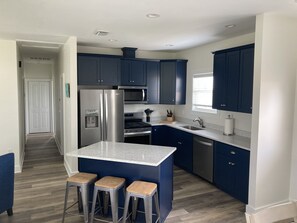 A modern kitchen with dark blue cabinets, a stainless steel refrigerator, a microwave, and a built-in oven. There are three stools at the island and a window providing natural light.