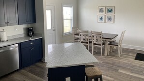 Una cocina con armarios azul oscuro, encimera blanca y cafetera. Hay una zona de comedor con mesa y sillas, y una ventana con persianas.