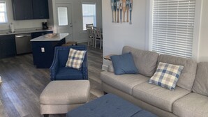 Una sala de estar moderna con sofá, reposapiés y sillón, una zona de cocina con armarios oscuros y un comedor con mesa y sillas.