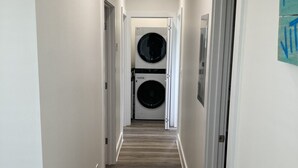 Un pasillo estrecho con suelo de madera, paredes blancas y un respiradero en el techo. Unos puertas dobles al final dan acceso a una sala de lavandería con dos lavadoras.
